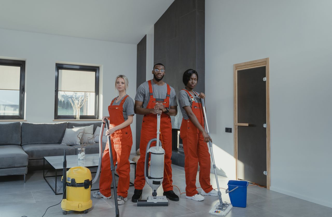 gallery-6 Diverse cleaning team in red uniforms ready to sanitize a modern living room with vacuum and mop.