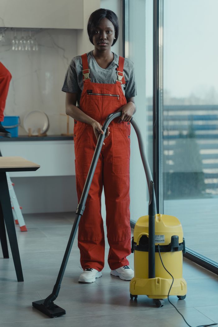 hero-img-02 An adult woman in red coveralls operates a vacuum cleaner in a modern indoor setting, focusing on cleaning tasks.